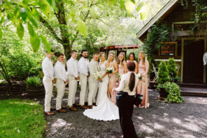 Snohomish Wedding Photographer GSquared Weddings A bride and groom stand with their wedding party, all dressed in light colors, posing for a photo outdoors near a wooden building surrounded by greenery. A photographer faces them, preparing to take the picture. Serving Seattle, Snohomish and Orlando