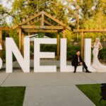 Snohomish Wedding Photographer GSquared Weddings A bride in a white gown and a groom in a black suit pose by large white letters spelling SNELL outdoors, while a photographer in black stands in the foreground. Trees and a wooden pergola are in the background. Serving Seattle, Snohomish and Orlando