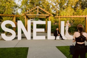 Snohomish Wedding Photographer GSquared Weddings A bride in a white gown and a groom in a black suit pose by large white letters spelling SNELL outdoors, while a photographer in black stands in the foreground. Trees and a wooden pergola are in the background. Serving Seattle, Snohomish and Orlando
