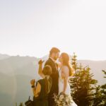 Snohomish Wedding Photographer GSquared Weddings A bride and groom stand close together outdoors with mountains in the background at sunset, while a woman adjusts the groom’s appearance. The bride holds a bouquet and wears a white dress. Serving Seattle, Snohomish and Orlando