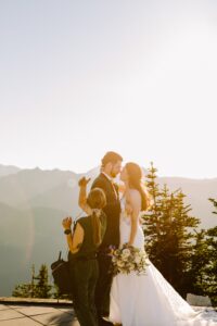 Snohomish Wedding Photographer GSquared Weddings A bride and groom stand close together outdoors with mountains in the background at sunset, while a woman adjusts the groom’s appearance. The bride holds a bouquet and wears a white dress. Serving Seattle, Snohomish and Orlando