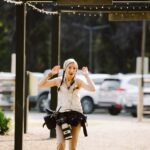 Snohomish Wedding Photographer GSquared Weddings A smiling woman with braided hair, wearing a hoodie and camera gear, raises her hands playfully while standing outdoors under string lights, with parked cars and greenery in the background. Serving Seattle, Snohomish and Orlando