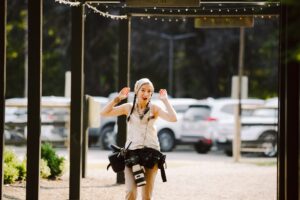 Snohomish Wedding Photographer GSquared Weddings A smiling woman with braided hair, wearing a hoodie and camera gear, raises her hands playfully while standing outdoors under string lights, with parked cars and greenery in the background. Serving Seattle, Snohomish and Orlando