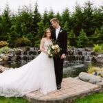 Snohomish Wedding Photographer GSquared Weddings A bride and groom stand together in front of a pond and garden. The bride holds a bouquet and wears a long white gown while an attendant adjusts her dress. The groom wears a black tuxedo and looks at the bride. Serving Seattle, Snohomish and Orlando