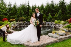 Snohomish Wedding Photographer GSquared Weddings A bride and groom stand together in front of a pond and garden. The bride holds a bouquet and wears a long white gown while an attendant adjusts her dress. The groom wears a black tuxedo and looks at the bride. Serving Seattle, Snohomish and Orlando