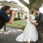 Snohomish Wedding Photographer GSquared Weddings A photographer takes a picture of a bride and groom standing close together, smiling, in front of a large tree and a house with a wraparound porch on a sunny day. Serving Seattle, Snohomish and Orlando