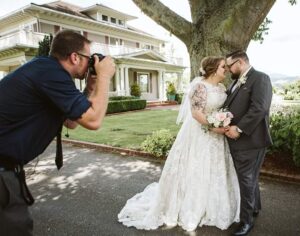 Snohomish Wedding Photographer GSquared Weddings A photographer takes a picture of a bride and groom standing close together, smiling, in front of a large tree and a house with a wraparound porch on a sunny day. Serving Seattle, Snohomish and Orlando