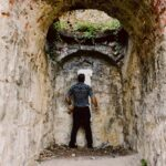 Snohomish Wedding Photographer GSquared Weddings A man stands with his back to the camera inside a narrow, arched stone tunnel, facing a rough stone wall with some greenery growing above it. Serving Seattle, Snohomish and Orlando