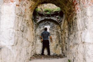 Snohomish Wedding Photographer GSquared Weddings A man stands with his back to the camera inside a narrow, arched stone tunnel, facing a rough stone wall with some greenery growing above it. Serving Seattle, Snohomish and Orlando