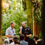 Snohomish Wedding Photographer GSquared Weddings Two men stand and talk under a flower-adorned arch in a lush outdoor setting, one holding a camera. They are surrounded by seated people, suggesting an event or ceremony, with greenery and soft lighting in the background. Serving Seattle, Snohomish and Orlando