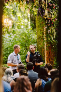 Snohomish Wedding Photographer GSquared Weddings Two men stand and talk under a flower-adorned arch in a lush outdoor setting, one holding a camera. They are surrounded by seated people, suggesting an event or ceremony, with greenery and soft lighting in the background. Serving Seattle, Snohomish and Orlando