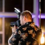 Snohomish Wedding Photographer GSquared Weddings A person wearing a black floral shirt is photographing an event indoors using a camera with a flash attachment. Bright lights are blurred in the background, creating a warm and vibrant atmosphere. Serving Seattle, Snohomish and Orlando