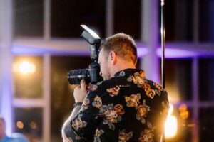 Snohomish Wedding Photographer GSquared Weddings A person wearing a black floral shirt is photographing an event indoors using a camera with a flash attachment. Bright lights are blurred in the background, creating a warm and vibrant atmosphere. Serving Seattle, Snohomish and Orlando