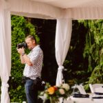 Snohomish Wedding Photographer GSquared Weddings A man in a patterned shirt stands under a white canopy, holding a camera and taking photos. There are white draped curtains, greenery in the background, and a decorated table with flowers and plates nearby. Serving Seattle, Snohomish and Orlando