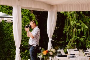 Snohomish Wedding Photographer GSquared Weddings A man in a patterned shirt stands under a white canopy, holding a camera and taking photos. There are white draped curtains, greenery in the background, and a decorated table with flowers and plates nearby. Serving Seattle, Snohomish and Orlando
