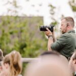 Snohomish Wedding Photographer GSquared Weddings A man in a green shirt with a headset uses a camera to film or take photos at an outdoor event, surrounded by a crowd of people. Trees and a building are in the blurred background. Serving Seattle, Snohomish and Orlando