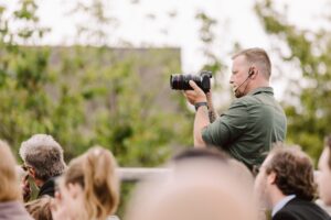 Snohomish Wedding Photographer GSquared Weddings A man in a green shirt with a headset uses a camera to film or take photos at an outdoor event, surrounded by a crowd of people. Trees and a building are in the blurred background. Serving Seattle, Snohomish and Orlando