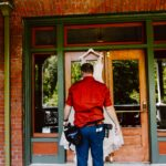 Snohomish Wedding Photographer GSquared Weddings A man in a red shirt and blue pants, holding camera equipment, stands in front of a wooden door, holding up a white wedding dress on a hanger against the glass. Serving Seattle, Snohomish and Orlando