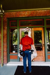 Snohomish Wedding Photographer GSquared Weddings A man in a red shirt and blue pants, holding camera equipment, stands in front of a wooden door, holding up a white wedding dress on a hanger against the glass. Serving Seattle, Snohomish and Orlando