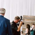 Snohomish Wedding Photographer GSquared Weddings A photographer in black clothing takes a picture at an indoor event with white drapes and string lights. Guests in formal attire are mingling in the background. Serving Seattle, Snohomish and Orlando