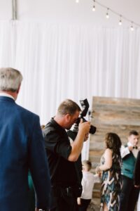 Snohomish Wedding Photographer GSquared Weddings A photographer in black clothing takes a picture at an indoor event with white drapes and string lights. Guests in formal attire are mingling in the background. Serving Seattle, Snohomish and Orlando