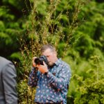 Snohomish Wedding Photographer GSquared Weddings A man wearing a blue, floral-patterned shirt and camera gear is photographing something outdoors, surrounded by green foliage and plants. Serving Seattle, Snohomish and Orlando