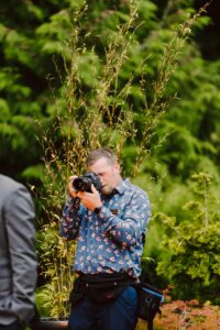 Snohomish Wedding Photographer GSquared Weddings A man wearing a blue, floral-patterned shirt and camera gear is photographing something outdoors, surrounded by green foliage and plants. Serving Seattle, Snohomish and Orlando