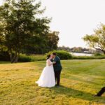 Snohomish Wedding Photographer GSquared Weddings A newlywed couple kisses on a sunlit grassy lawn near a lake, surrounded by trees, while a photographer kneels nearby to capture the moment. Serving Seattle, Snohomish and Orlando