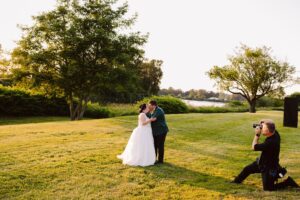 Snohomish Wedding Photographer GSquared Weddings A newlywed couple kisses on a sunlit grassy lawn near a lake, surrounded by trees, while a photographer kneels nearby to capture the moment. Serving Seattle, Snohomish and Orlando