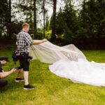 Snohomish Wedding Photographer GSquared Weddings A photographer crouches to take a picture while an assistant holds up the bride’s long veil outdoors on a grassy area surrounded by tall trees. The bride sits in a white dress holding a bouquet. Serving Seattle, Snohomish and Orlando