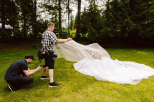 Snohomish Wedding Photographer GSquared Weddings A photographer crouches to take a picture while an assistant holds up the bride’s long veil outdoors on a grassy area surrounded by tall trees. The bride sits in a white dress holding a bouquet. Serving Seattle, Snohomish and Orlando