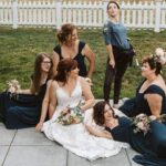 Snohomish Wedding Photographer GSquared Weddings A bride in a white gown sits on the ground surrounded by bridesmaids in dark blue dresses, holding bouquets, while a casually dressed woman stands behind them, posing playfully. They are outside near a white picket fence. Serving Seattle, Snohomish and Orlando