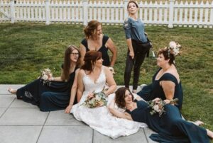 Snohomish Wedding Photographer GSquared Weddings A bride in a white gown sits on the ground surrounded by bridesmaids in dark blue dresses, holding bouquets, while a casually dressed woman stands behind them, posing playfully. They are outside near a white picket fence. Serving Seattle, Snohomish and Orlando