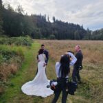 Snohomish Wedding Photographer GSquared Weddings A bride and groom pose together on a grassy path in a field, surrounded by trees, while two photographers capture their wedding photos under a cloudy sky. Serving Seattle, Snohomish and Orlando
