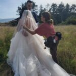 Snohomish Wedding Photographer GSquared Weddings A bride and groom stand outdoors in a grassy field with trees in the background, while a woman adjusts the brides flowing wedding dress and veil. Serving Seattle, Snohomish and Orlando