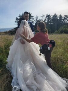 Snohomish Wedding Photographer GSquared Weddings A bride and groom stand outdoors in a grassy field with trees in the background, while a woman adjusts the brides flowing wedding dress and veil. Serving Seattle, Snohomish and Orlando