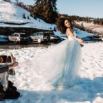 Snohomish Wedding Photographer GSquared Weddings A woman in a flowing blue dress poses in the snow while two people adjust her dress and take photos. Cars are parked nearby, and snowy hills and trees are in the background under a clear sky. Serving Seattle, Snohomish and Orlando