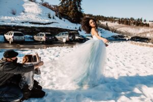Snohomish Wedding Photographer GSquared Weddings A woman in a flowing blue dress poses in the snow while two people adjust her dress and take photos. Cars are parked nearby, and snowy hills and trees are in the background under a clear sky. Serving Seattle, Snohomish and Orlando