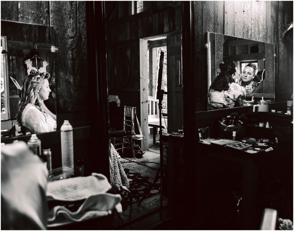 Snohomish Wedding Photographer GSquared Weddings In a rustic Snohomish room, a bride sits before a mirror, preparing for her wedding. The reflection shows her smiling face amid wedding essentials. An open door leads outside as natural light filters through the space. Black and white photo. Serving Seattle, Snohomish and Orlando