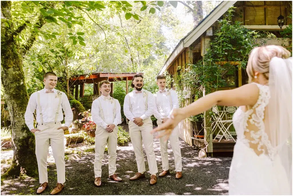 Snohomish Wedding Photographer GSquared Weddings A bride, seen from behind, approaches four groomsmen in matching beige pants and white shirts, all smiling at her. They stand under lush green trees next to a rustic wooden building on a sunny Snohomish day. Serving Seattle, Snohomish and Orlando