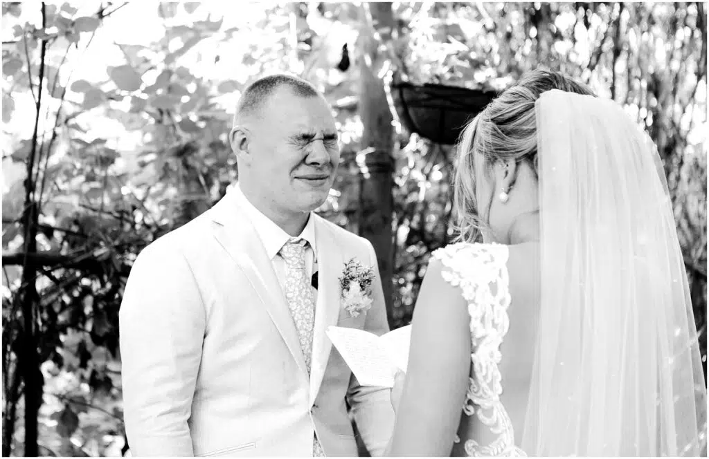 Snohomish Wedding Photographer GSquared Weddings Amidst the lush greenery of Snohomish, a bride and groom stand during their wedding ceremony. The groom, in a light suit, appears emotional as the bride, wearing a lacy dress and veil, reads from a paper. Capturing this moment in black and white adds to its timeless elegance. Serving Seattle, Snohomish and Orlando