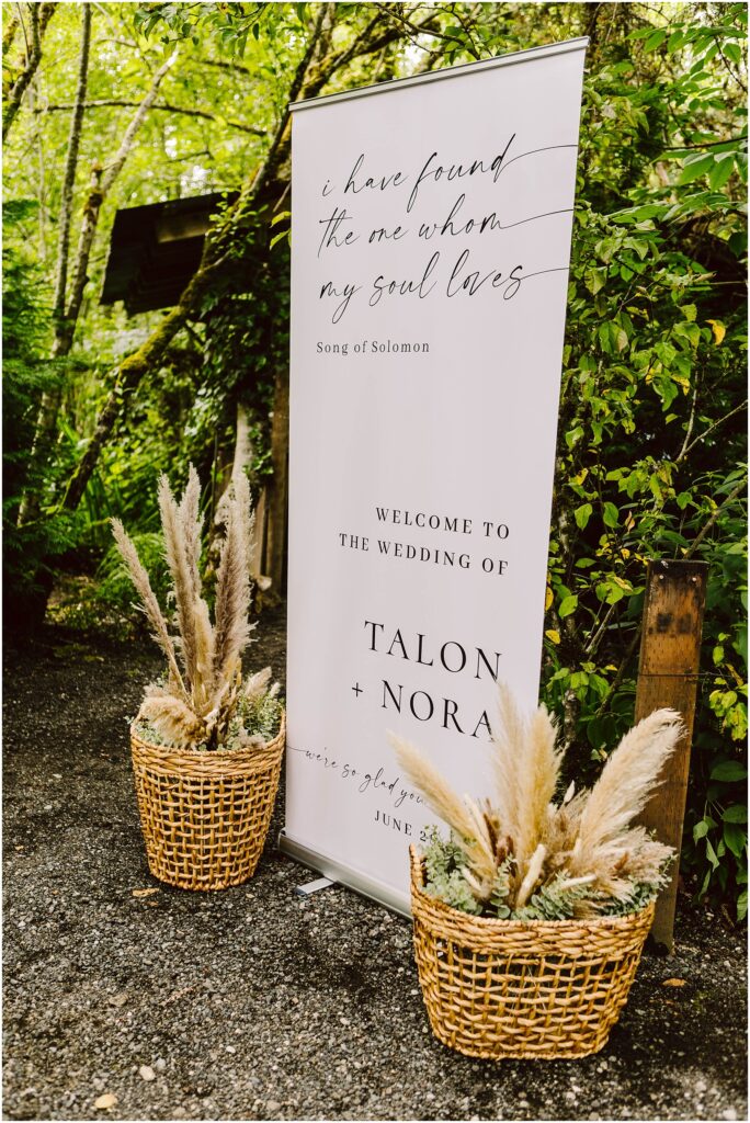 Snohomish Wedding Photographer GSquared Weddings In a picturesque Snohomish garden, a wedding sign adorned with lush greenery welcomes guests to the Wedding of Talon + Nora. A quote from Song of Solomon graces the sign, while two baskets filled with pampas grass enhance its charming allure. Serving Seattle, Snohomish and Orlando