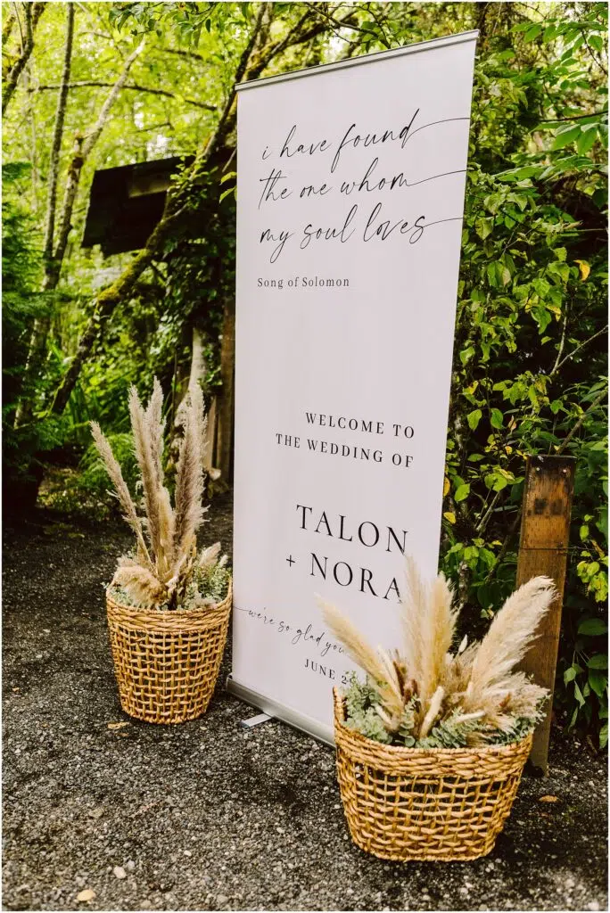 Snohomish Wedding Photographer GSquared Weddings In a picturesque Snohomish garden, a wedding sign adorned with lush greenery welcomes guests to the Wedding of Talon + Nora. A quote from Song of Solomon graces the sign, while two baskets filled with pampas grass enhance its charming allure. Serving Seattle, Snohomish and Orlando