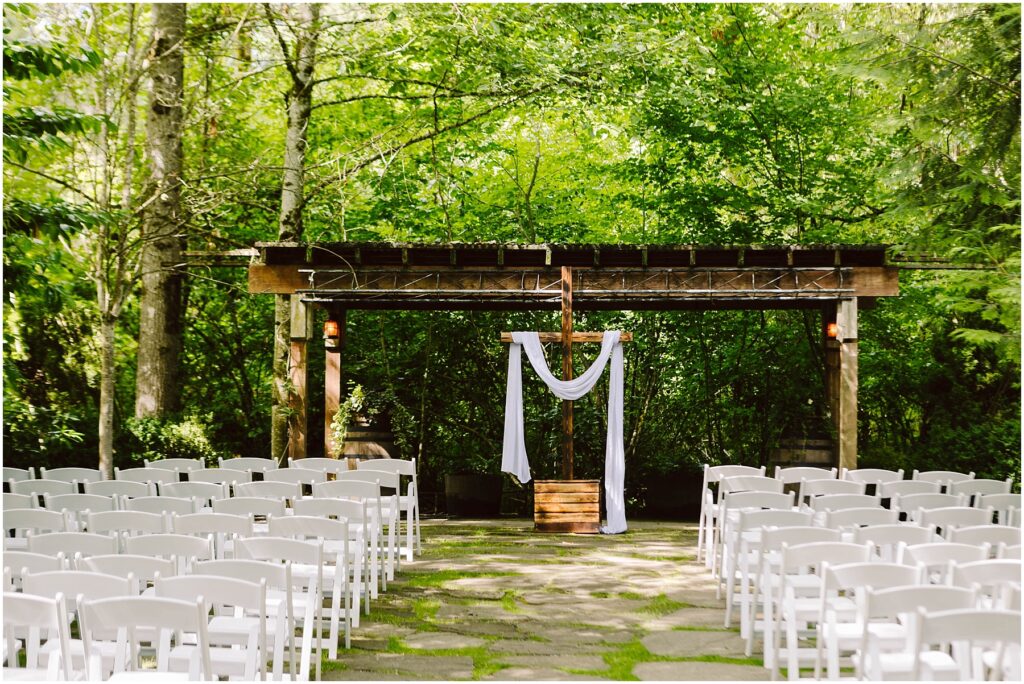 Snohomish Wedding Photographer GSquared Weddings An outdoor wedding setup in Snohomish features rows of white chairs facing a wooden arbor draped with white fabric. The lush green trees and foliage create a serene forest setting under clear daylight, perfect for a tranquil celebration. Serving Seattle, Snohomish and Orlando