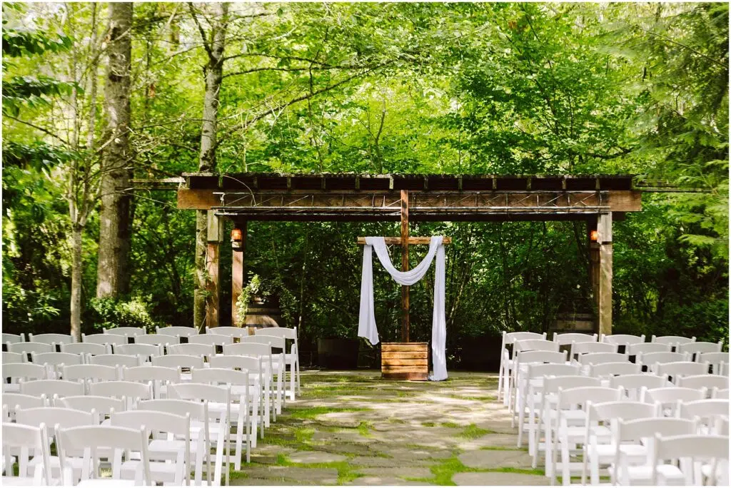 Snohomish Wedding Photographer GSquared Weddings An outdoor wedding setup in Snohomish features rows of white chairs facing a wooden arbor draped with white fabric. The lush green trees and foliage create a serene forest setting under clear daylight, perfect for a tranquil celebration. Serving Seattle, Snohomish and Orlando
