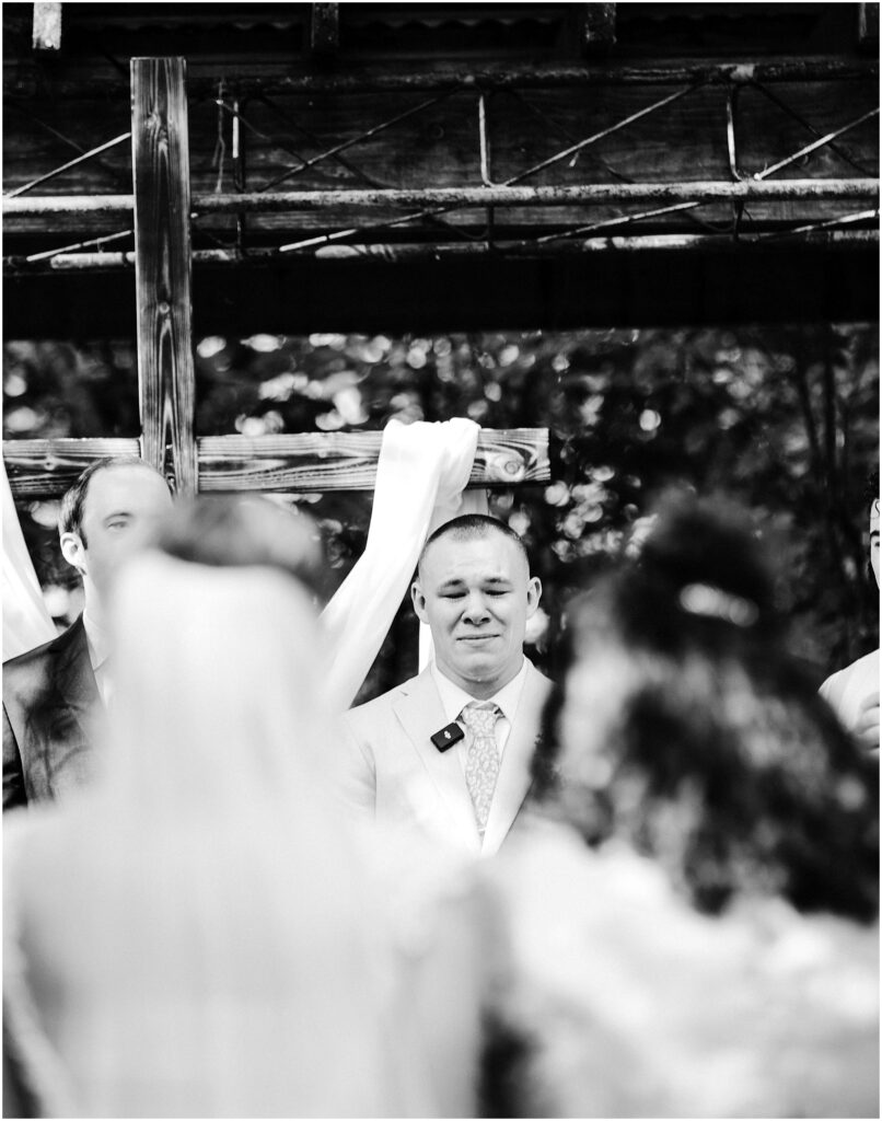 Snohomish Wedding Photographer GSquared Weddings A groom in a light-colored suit stands at an outdoor altar during a wedding ceremony in Snohomish. He looks toward the bride, who is slightly blurred in the foreground, as trees and wooden beams beautifully frame the romantic setting. Serving Seattle, Snohomish and Orlando
