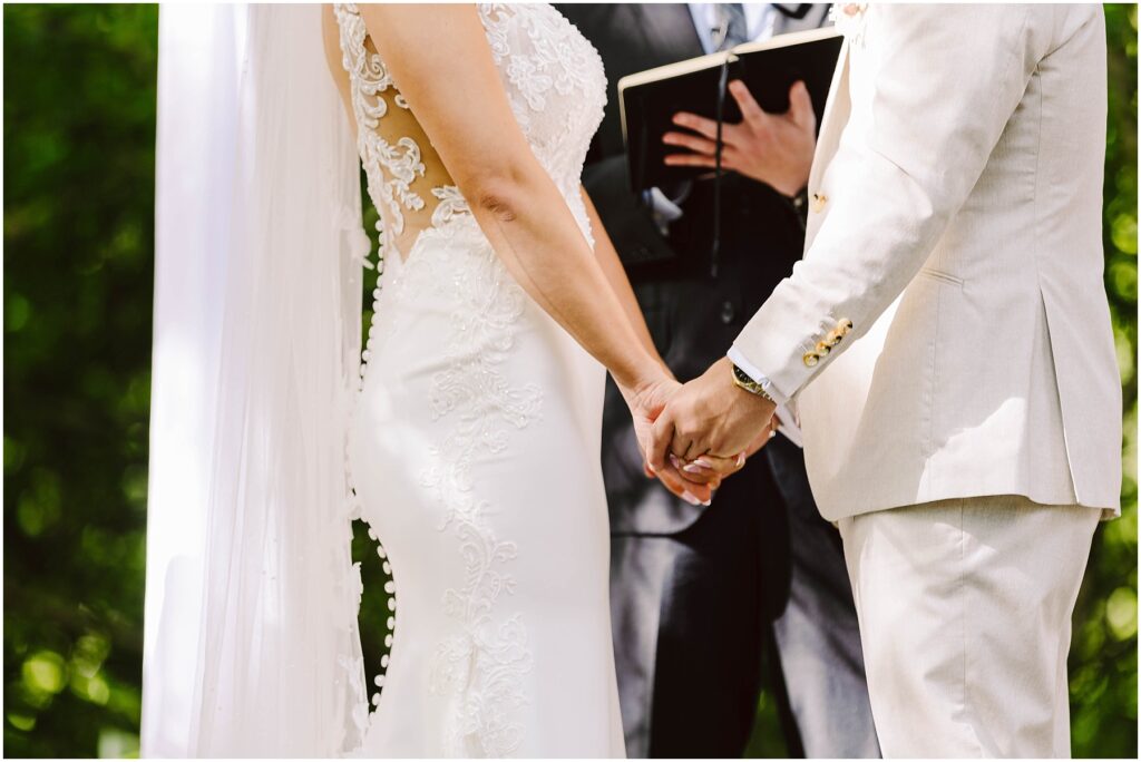 Snohomish Wedding Photographer GSquared Weddings A bride in a lace dress and a groom in a cream suit stand hand-in-hand during an outdoor wedding ceremony in Snohomish. A person in a suit stands behind them with an open book, likely officiating the ceremony. Serving Seattle, Snohomish and Orlando