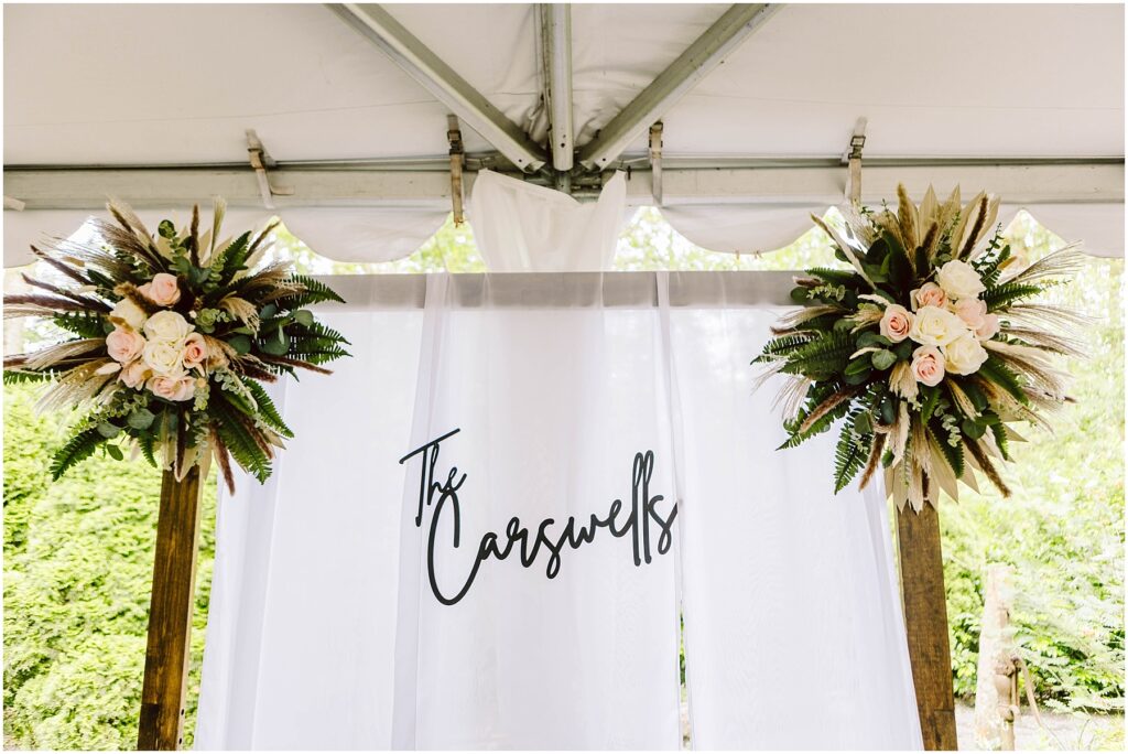 Snohomish Wedding Photographer GSquared Weddings A white wedding backdrop with the text The Carswells in elegant script stands gracefully in a Snohomish setting. Flanking the backdrop are two floral arrangements featuring pink roses, greenery, and dried palm leaves, all under a tent. Serving Seattle, Snohomish and Orlando
