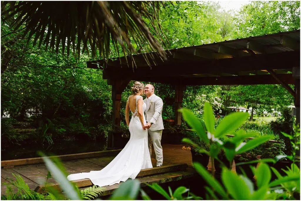 Snohomish Wedding Photographer GSquared Weddings A bride and groom stand together under a wooden gazebo in Snohomish, surrounded by lush greenery. The bride wears a long white dress with a flowing train, while the groom is in a light-colored suit. They hold hands and gaze at each other lovingly. Serving Seattle, Snohomish and Orlando