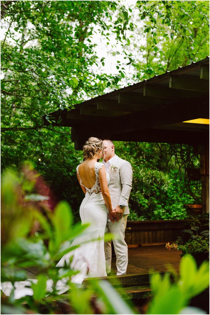 Snohomish Wedding Photographer GSquared Weddings In a lush, green garden setting in Snohomish, a bride and groom share a tender kiss. The bride dazzles in her lace-detailed dress while the groom stands beside her in a light-colored suit on a wooden platform, embraced by vibrant foliage. Serving Seattle, Snohomish and Orlando